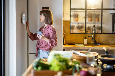 Woman using digital tablet hanging on the wall in the kitchen, controlling smart home devices. Modern kitchen interior with food in frontの写真素材