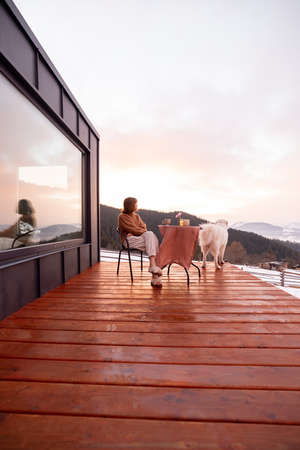 Woman with her dog resting on terrace of tiny house in the mountains, enjoying beautiful landscape during sunrise. Concept of small modern cabins for rest and escape to natureの写真素材