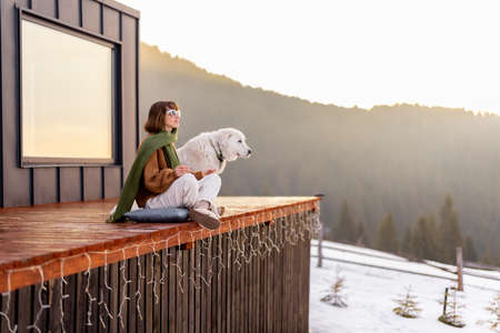 Woman sitting with dog on terrace of tiny house in the mountains. Concept of small modern cabins for rest and escape to nature. Idea of traveling with dogの写真素材