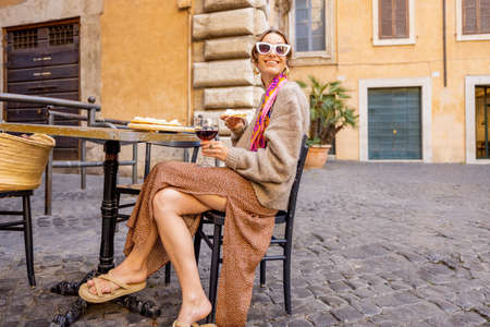 Woman eating pizza and drinking wine at restaurant on a street in Rome. Concept of Italian gastronomy and travel. Stylish woman with sunglasses and colorful hair shawlの写真素材