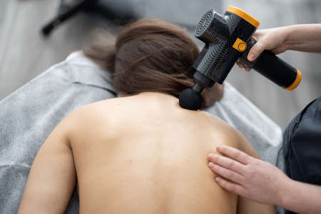 Woman massages the patients back with a percussion massager. Concept of new technologies for acupressure of trigger points. Therapeutic massage and back treatmentの写真素材