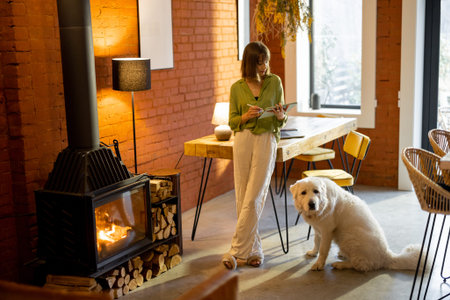 Young woman works on digital tablet while standing with her adorable dog at cozy living room....