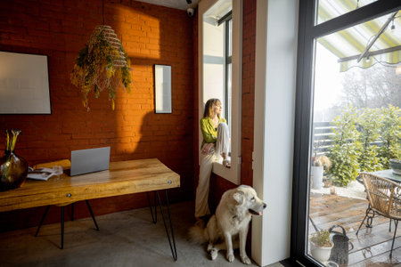 Woman sitting on the window sill and looking out the window with her white adorable dog at home....
