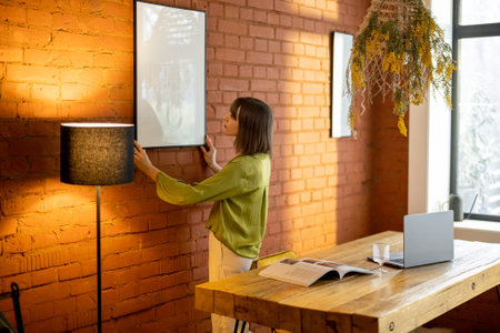 Young woman hangs and fixes photo frame on brick wall, decorating living space at home. Mockup image...