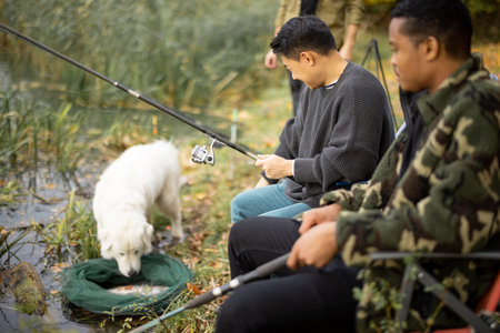 Multiracial male friends fishing with dog on river or lake coast. Concept of leisure, hobby and...