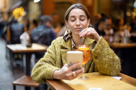 Young woman using phone while sitting with spritz aperol drink at bar outdoors. Concept of Italian lifestyle and gastronomy. Idea of remote online communicationの写真素材