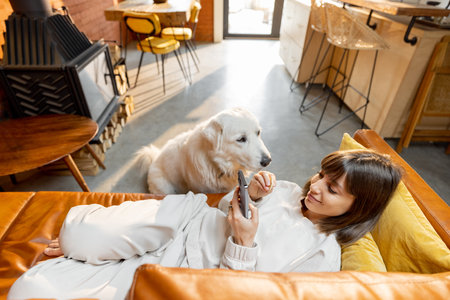 Young woman lying relaxed on a couch and using phone, spending time with her cute adorable dog at...