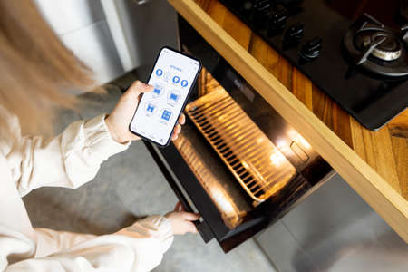 Woman holding phone with running smart home application, controlling kitchen devices remotely. Smart kitchen appliances conceptの写真素材