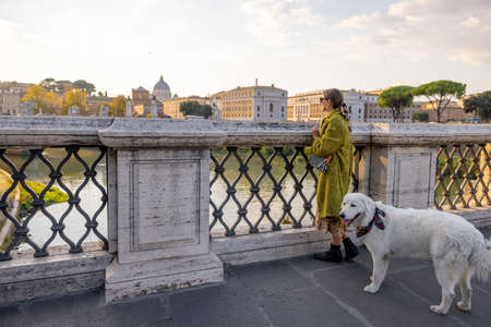Woman enjoying beautiful view of Rome city, standing with her dog on the bridge during the morning time. Elegant woman wearing coat and shawl in hair. Concept of italian lifestyle and travelの写真素材