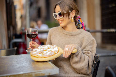 Woman eating pizza and drinking wine at restaurant on a street in Rome. Concept of Italian gastronomy and travel. Stylish woman with sunglasses and colorful hair shawlの写真素材