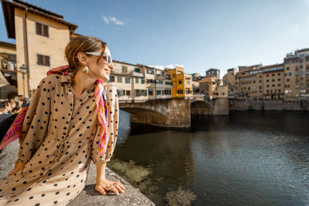 Young woman enjoys beautiful view on famous Old bridge in Florence, sitting on the riverside at sunset. Female traveler visiting italian landmarks. Stylish woman wearing dress and colorful shawlの写真素材