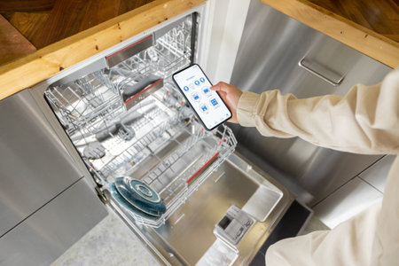 Woman holding phone with running smart home application, controlling dishwasher remotely. Smart kitchen appliances conceptの写真素材