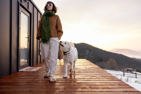 Woman walks with her dog on terrace of tiny house in the mountains during winter time. Concept of...