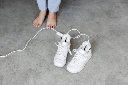 Woman stands barefoot near white sneakers drying with electric shoe dryer on the floor. Concept of...