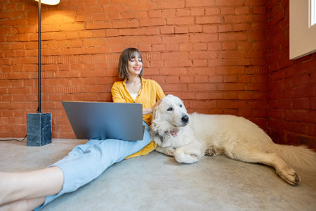 Young woman working on laptop computer while sitting with her cute dog on brick wall background at...
