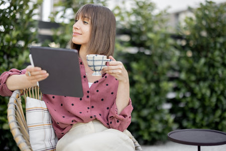 Young creative woman works on digital tablet while sitting relaxed with coffee cup at backyard of...