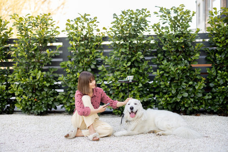 Young woman sitting relaxed with her huge white adorable dog on green hedge background, spending...