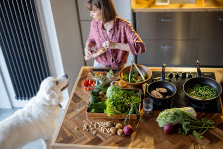 Young housewife makes a salad while standing with her adorable white dog in the kitchen. Table with lots of fresh green food ingredients. Healthy eating and leisure time with petの写真素材
