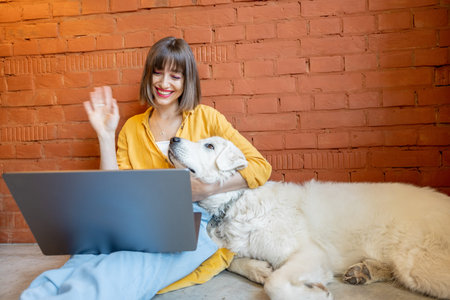 Young cheerful woman having video call on laptop computer while sitting with her cute white dog on...