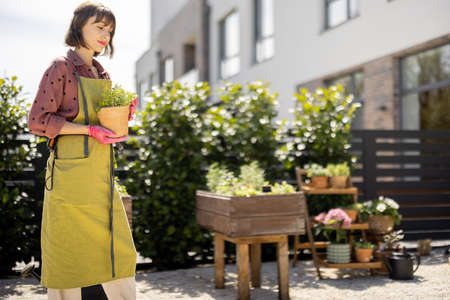 Portrait of young and cheerful gardener in green apron holding pot with herbs at home vegetable garden in backyard. Concept of hobby, homegrowing and leisure timeの写真素材
