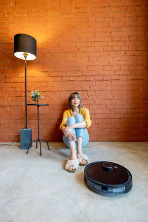 Young woman sitting relaxed on floor while robotic vacuum cleaner cleaning floor at home. Concept of smart home gadgets and comfortの写真素材