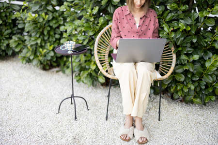 Young woman works on laptop computer while sitting relaxed on chair on background of green bushes at backyard. Concept of remote work at cozy atmosphereの写真素材