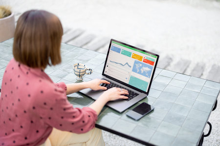 Young woman works on laptop while sitting by the table outdoors. View from the backside on computer...