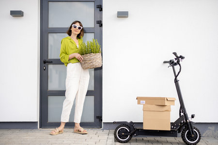 Young woman carrying basket with herbs on a porch of her house with electric scooter and parcels...