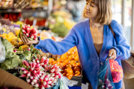 Woman takes a bunch of radishes from the counter, buying fresh vegetables and fruits at the local market. Shopping with reusable mesh bag. Sustainability and organic food conceptの写真素材