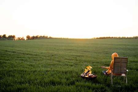 Young woman in casual overalls sitting by the fireplace and enjoying beautiful sunset on green field alone. Wide view with copy space. Escaping to nature and summer recreation conceptの写真素材
