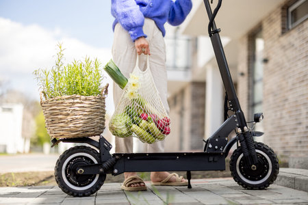 Woman holds mesh bag full of fresh vegetables going to drive on electrical scooter with with rosemary in a basket on trunk, cropped view. Concept of eco-friendly amd modern lifestyleの写真素材