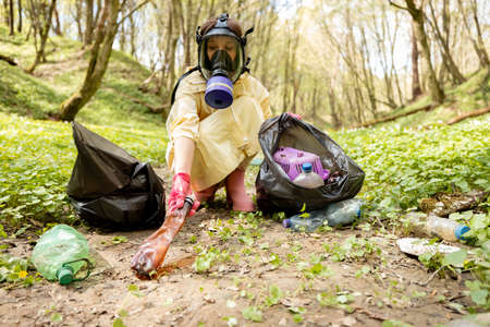 Woman in gas mask and protective clothes collecting scattered plastic garbage in the woodsの写真素材