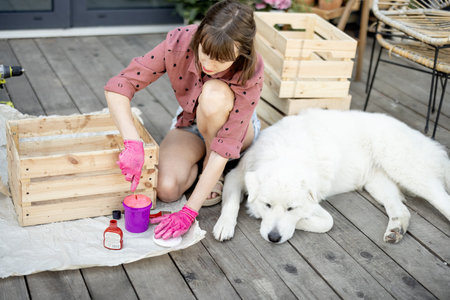 Young woman painting wooden box in pink color, doing housework while sitting with her dog on terrace...