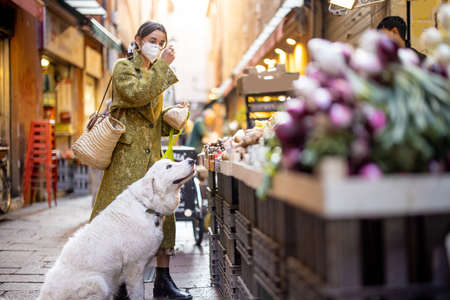 Woman in medical mask buying fruits and vegetables with dog at market stall on the famous gastronomical street in Bologna. Concept of buying local products during pandemic. Idea of Italian lifestyleの写真素材