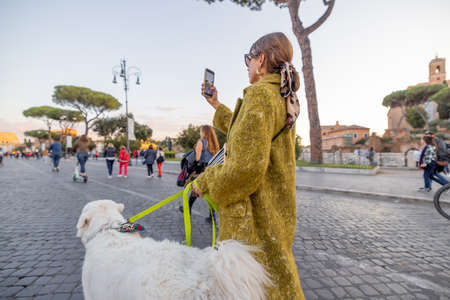 Woman walking with a dog on the famous central street in Rome, taking photos on phone and enjoying old architecture during a sunset. Concept of italian lifestyle and travelの写真素材