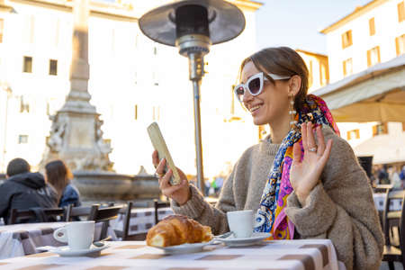 Woman having video call on smart phone while sitting at traditional italian cafe outdoors. Idea of spending time in Rome. Concept of italian lifestyle and travelの写真素材
