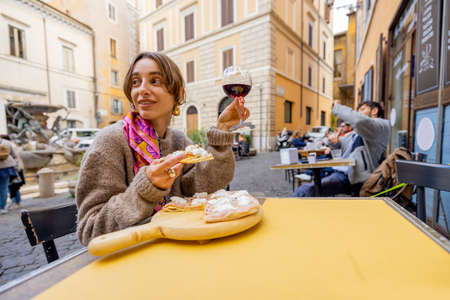 Woman eating pizza and drinking wine at restaurant on a street in Rome. Concept of Italian gastronomy and travel. Stylish woman with sunglasses and colorful hair shawlの写真素材