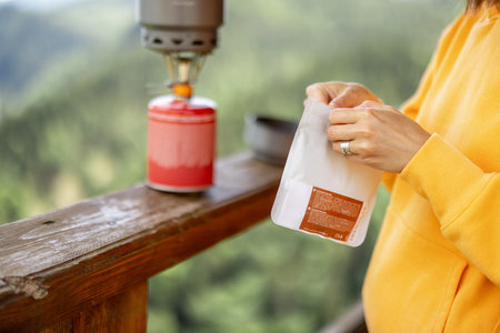 Young woman prepares freeze-dried food for hiking in airtight package, with gas burner, while...