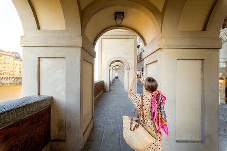 Young woman photographing on phone beautiful archway, visiting famous italian city Florence. Female tourist enjoys italian old architecture. Woman dressed in Italian style with colorful scarf in hairの写真素材