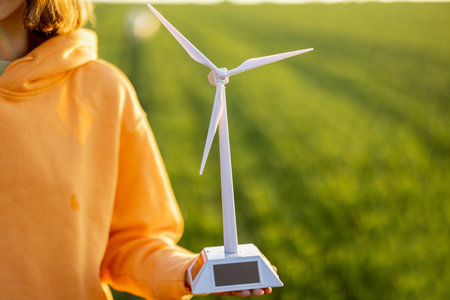 Person holds toy wind generator on green field background on sunset, close-up. Concept of ecological...