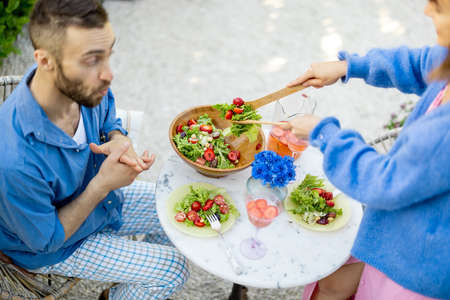 Excited man is waiting for lunch, watching enthusiastically as his wife serves healthy salad. Young family have a lunch at backyard during summer timeの写真素材