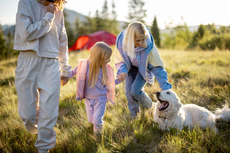 Two women walk with little girl and white dog, happily spending summer time on nature. Homosexual family with a kid travel in mountainsの写真素材
