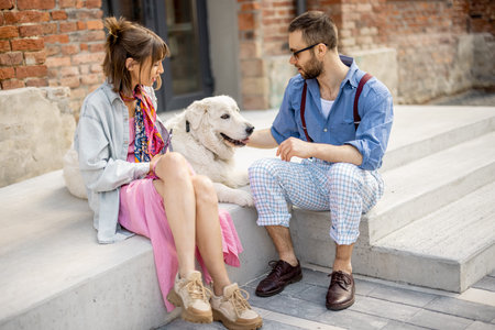 Young stylish couple sit together and have fun with their white dog on a street. Young hipsters hang...