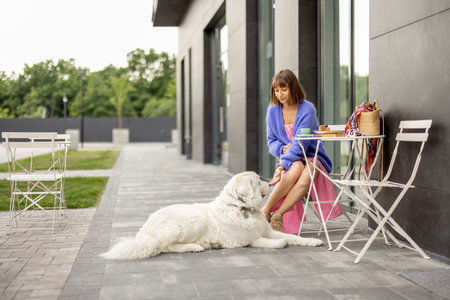 Young stylish woman sits with her cute adorable dog at cafe on a street. Leisure time with pet...