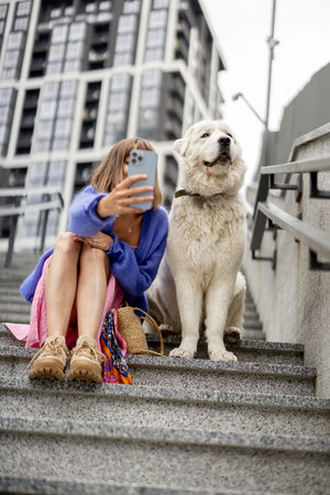 Cheerful woman takes selfie on phone with her dog while sitting on staircase at modern residential...