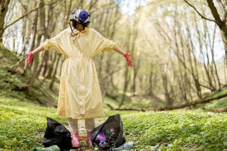Woman in gas mask and protective clothes stands with garbage bags collecting scattered plastic in the woods. Problem of environmental pollution. Safe the world conceptの写真素材