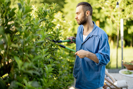 Stylish guy pruning green bushes with scissors at garden. Handsome gardener during job outdoors
