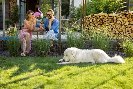 Young stylish couple have a drink while sitting together with dog on a lawn at their beautiful backyard of country house. Young man and woman spend summer timeの写真素材