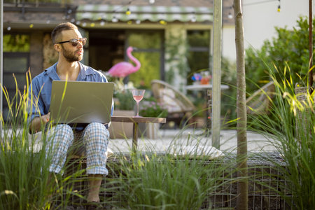 Cool guy works on laptop while sitting on porch of his country house. Concept of remote work at cozy home workplace and freelanceの写真素材