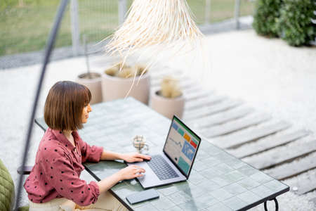 Young woman works on laptop while sitting by the table outdoors. View from the backside on computer screen with charts. Concept of remote online workの写真素材